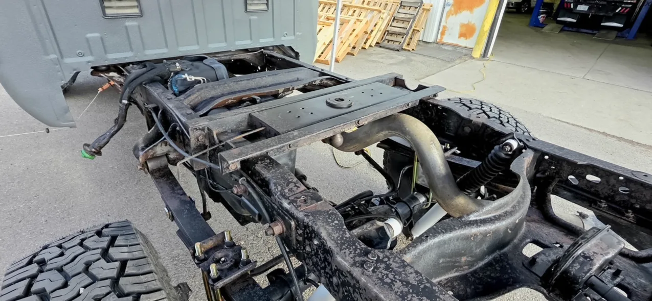 Overhead view of a pickup chassis with the bed removed, showing frame rails, crossmembers, exhaust routing and suspension components at a shop.