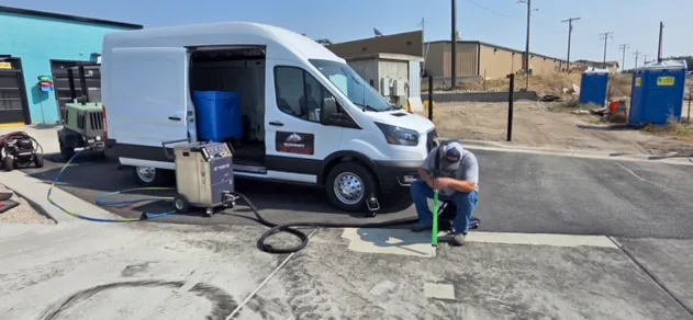 Summit Cryo technician kneeling with a blast nozzle on a concrete pad beside the mobile rig van, hoses running back to a towable diesel compressor.