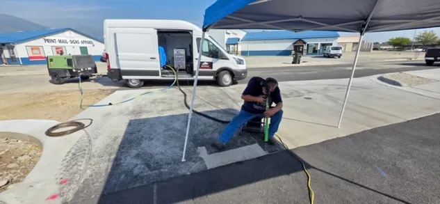 Technician blasting a concrete surface under a pop-up canopy, with the rig van and diesel compressor parked behind and blast hose running across the lot.