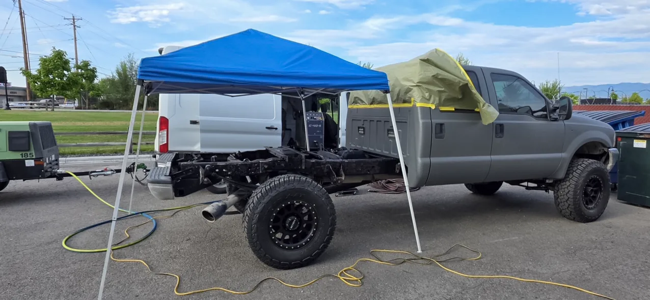 Pickup truck with the bed removed and its chassis exposed, parked between a work canopy and the mobile rig van with blast hoses laid out on the pavement.